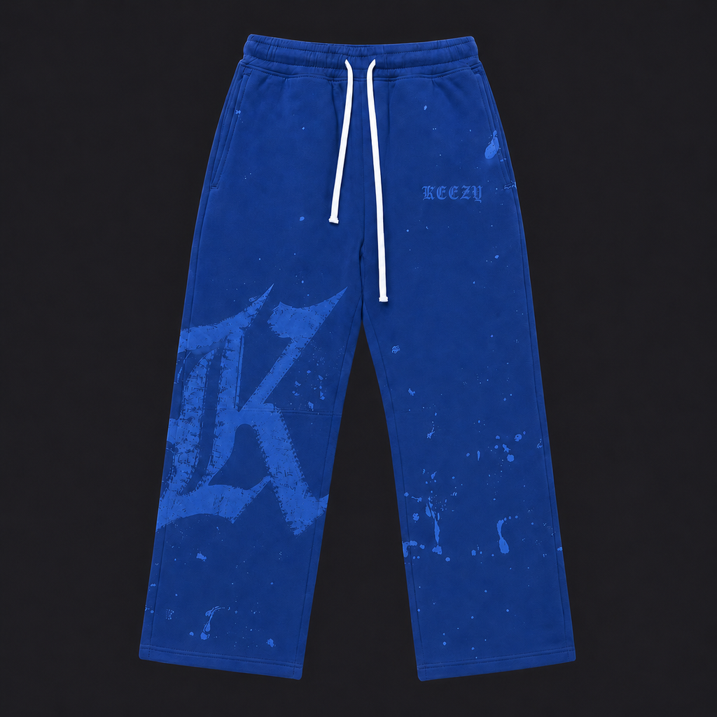 Blue sweatpants with a white drawstring, featuring a large light blue distressed 'K' graphic on the left leg, a small 'keezy' logo on the right thigh, and paint splatter details, shown against a black background.