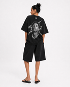 A model shown from the back wearing a black oversized t-shirt with a large graphic print of a face and starburst design, paired with black shorts and black sandals, against a plain white studio background. FOOTBALL. JUDE BELLINGHAM