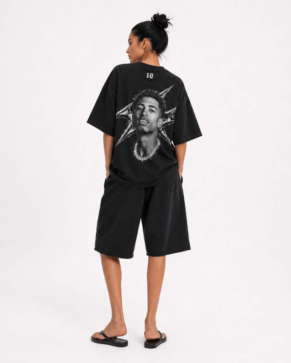 A model shown from the back wearing a black oversized t-shirt with a large graphic print of a face and starburst design, paired with black shorts and black sandals, against a plain white studio background. FOOTBALL. JUDE BELLINGHAM