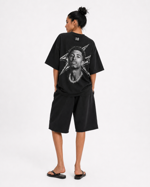 A model shown from the back wearing a black oversized t-shirt with a large graphic print of a face and starburst design, paired with black shorts and black sandals, against a plain white studio background. FOOTBALL. JUDE BELLINGHAM