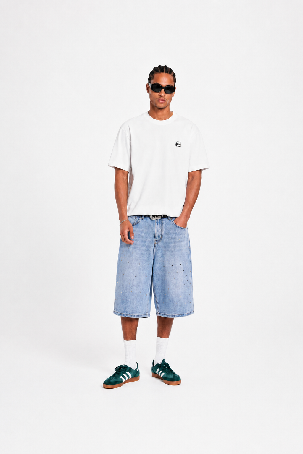 A man modeling a white short-sleeve t-shirt with a small black chest logo, light-wash denim shorts, white socks, and green sneakers, standing against a plain white studio background.