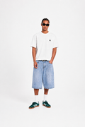 A man modeling a white short-sleeve t-shirt with a small black chest logo, light-wash denim shorts, white socks, and green sneakers, standing against a plain white studio background.