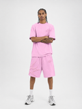 A man modeling a matching light pink lounge set consisting of a short-sleeve t-shirt and distressed drawstring shorts, paired with white socks and gray sneakers, against a plain light gray background.