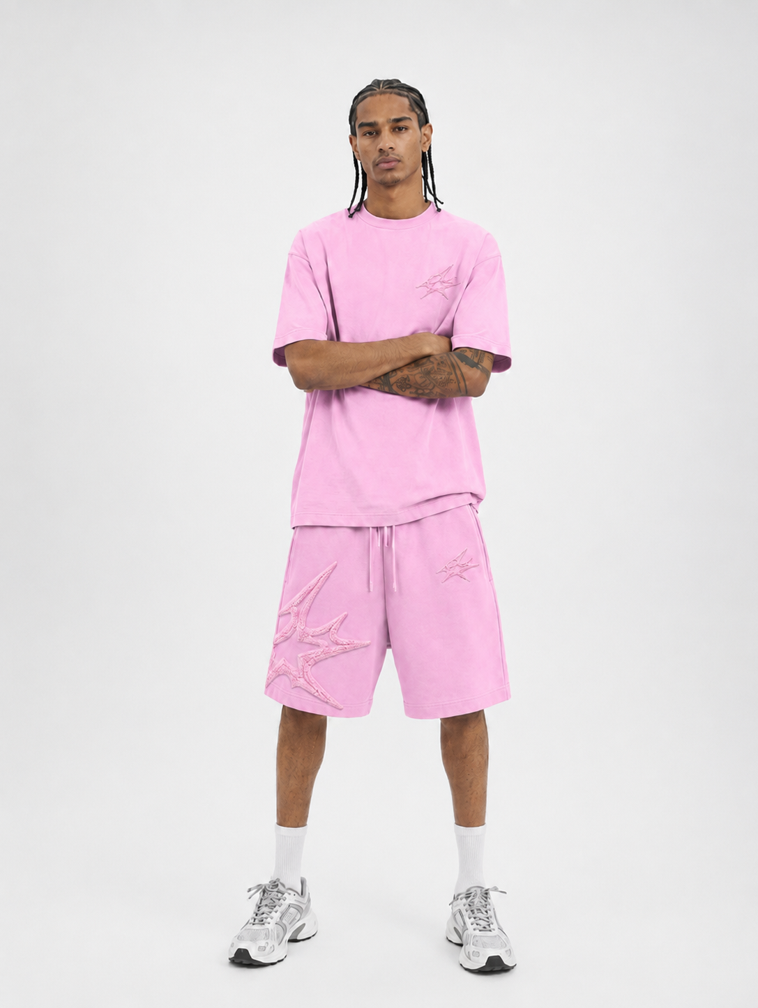 A man modeling a matching light pink lounge set consisting of a short-sleeve t-shirt and distressed drawstring shorts, paired with white socks and gray sneakers, against a plain light gray background.