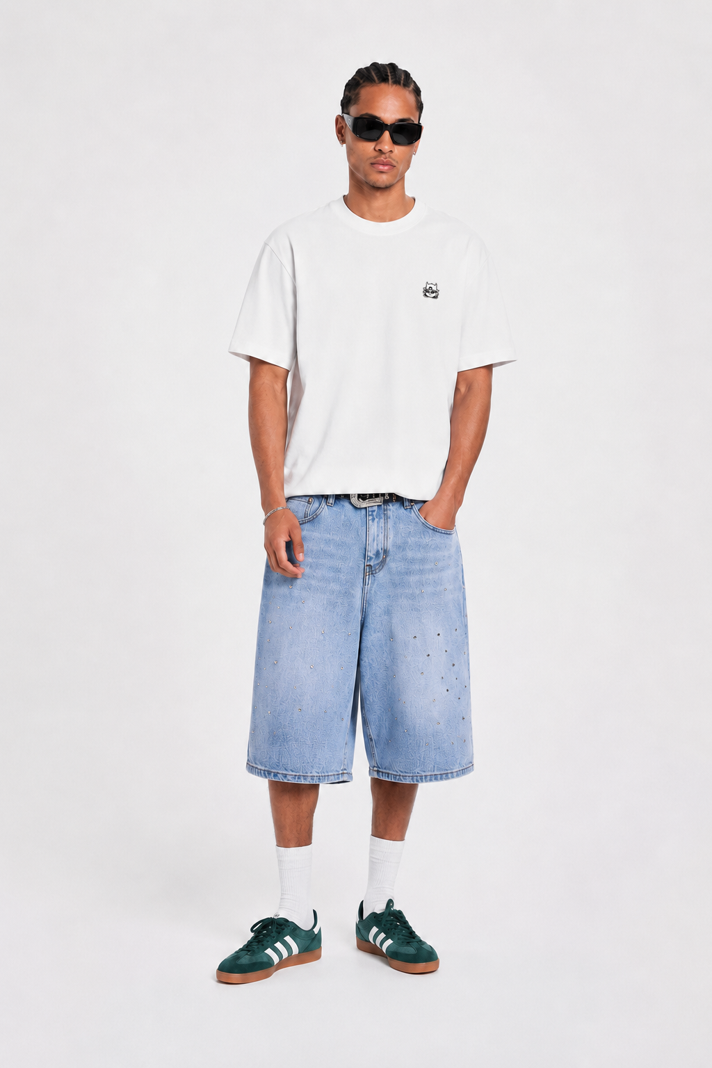 A man modeling a white short-sleeve t-shirt with a small black chest logo, light-wash denim shorts, white socks, and green sneakers, standing against a plain light gray studio background.