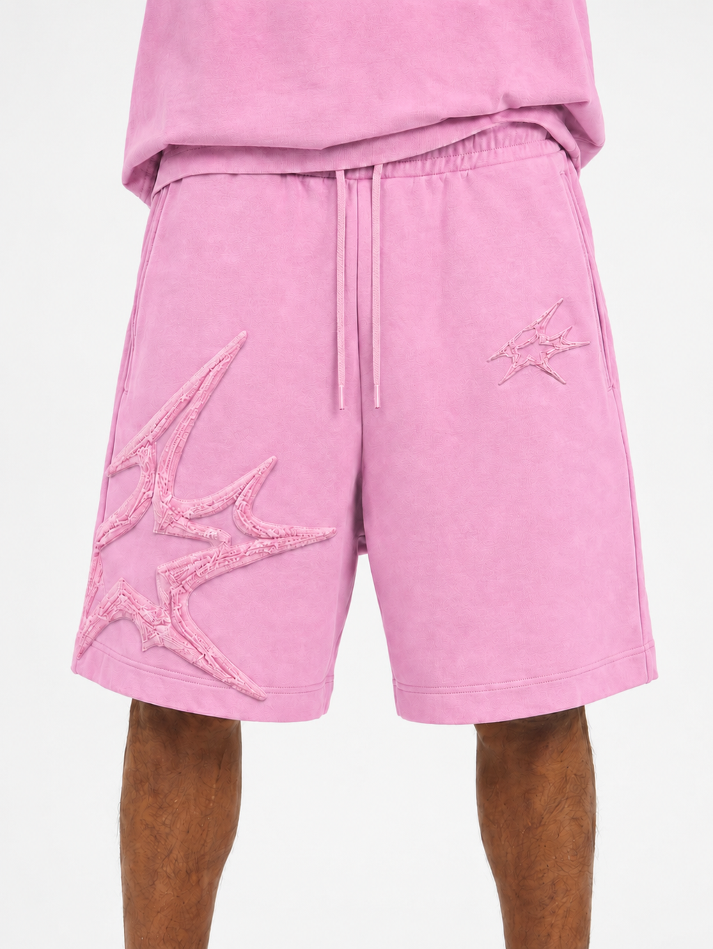 Pink athletic shorts with a drawstring waist and large tonal embroidered starburst graphics on the left leg, shown on a model against a light gray background.