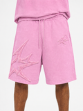 Pink athletic shorts with a drawstring waist and large tonal embroidered starburst graphics on the left leg, shown on a model against a light gray background.