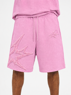 Pink athletic shorts with a drawstring waist and large tonal embroidered starburst graphics on the left leg, shown on a model against a light gray background.