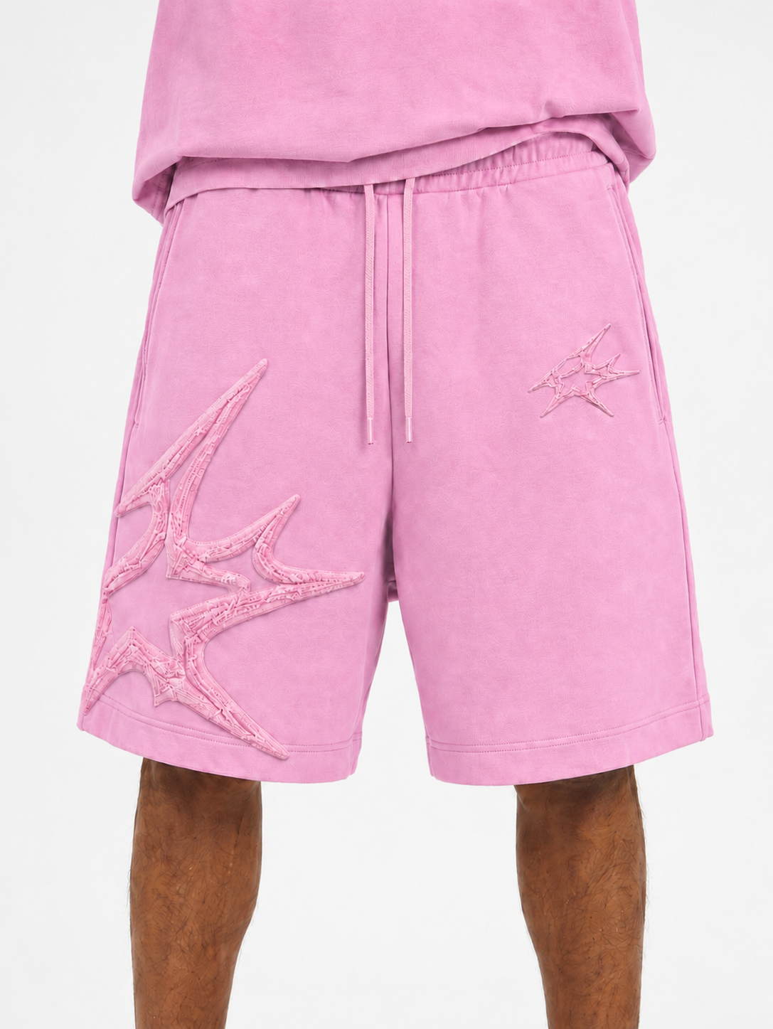 Pink athletic shorts with a drawstring waist and large tonal embroidered starburst graphics on the left leg, shown on a model against a light gray background.