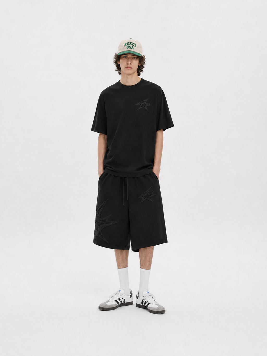 A model stands against a light gray studio background wearing a black short-sleeve t-shirt with a subtle tonal star graphic on the chest, matching black shorts, white crew socks, and white sneakers. He is also wearing a beige baseball cap with green and black lettering.