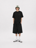 A model stands against a light gray studio background wearing a black short-sleeve t-shirt with a subtle tonal star graphic on the chest, matching black shorts, white crew socks, and white sneakers. He is also wearing a beige baseball cap with green and black lettering.