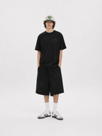 A model stands against a light gray studio background wearing a black short-sleeve t-shirt with a subtle tonal star graphic on the chest, matching black shorts, white crew socks, and white sneakers. He is also wearing a beige baseball cap with green and black lettering.