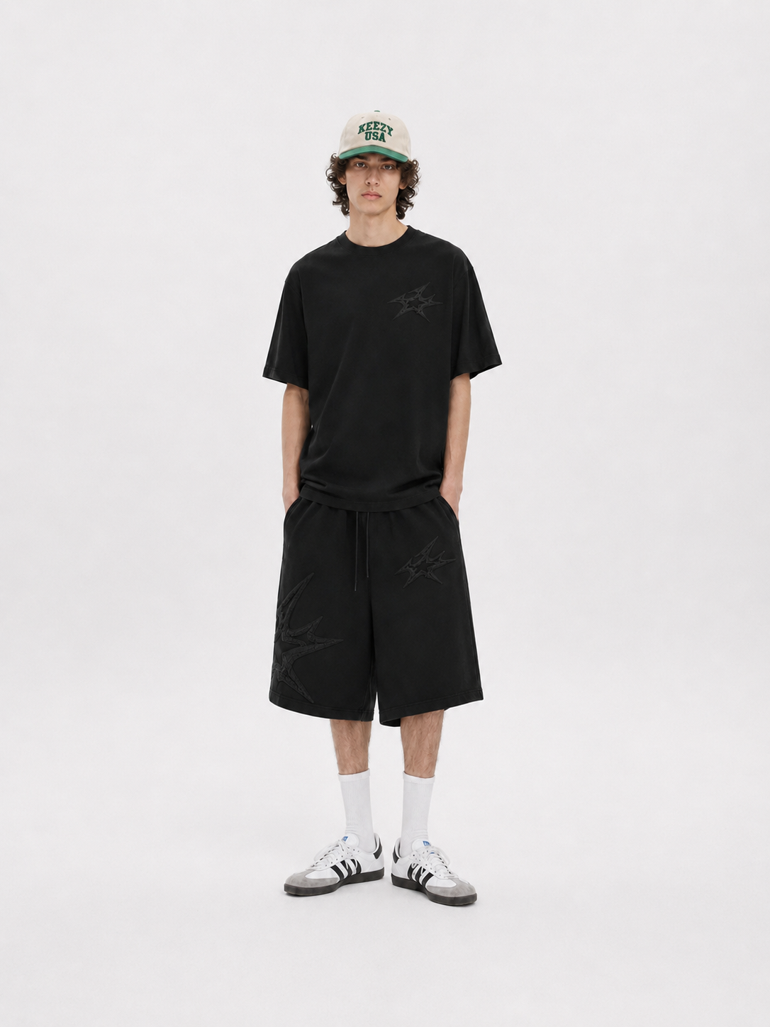 A model stands against a light gray studio background wearing a black short-sleeve t-shirt with a subtle tonal star graphic on the chest, matching black shorts, white crew socks, and white sneakers. He is also wearing a beige baseball cap with green and black lettering.