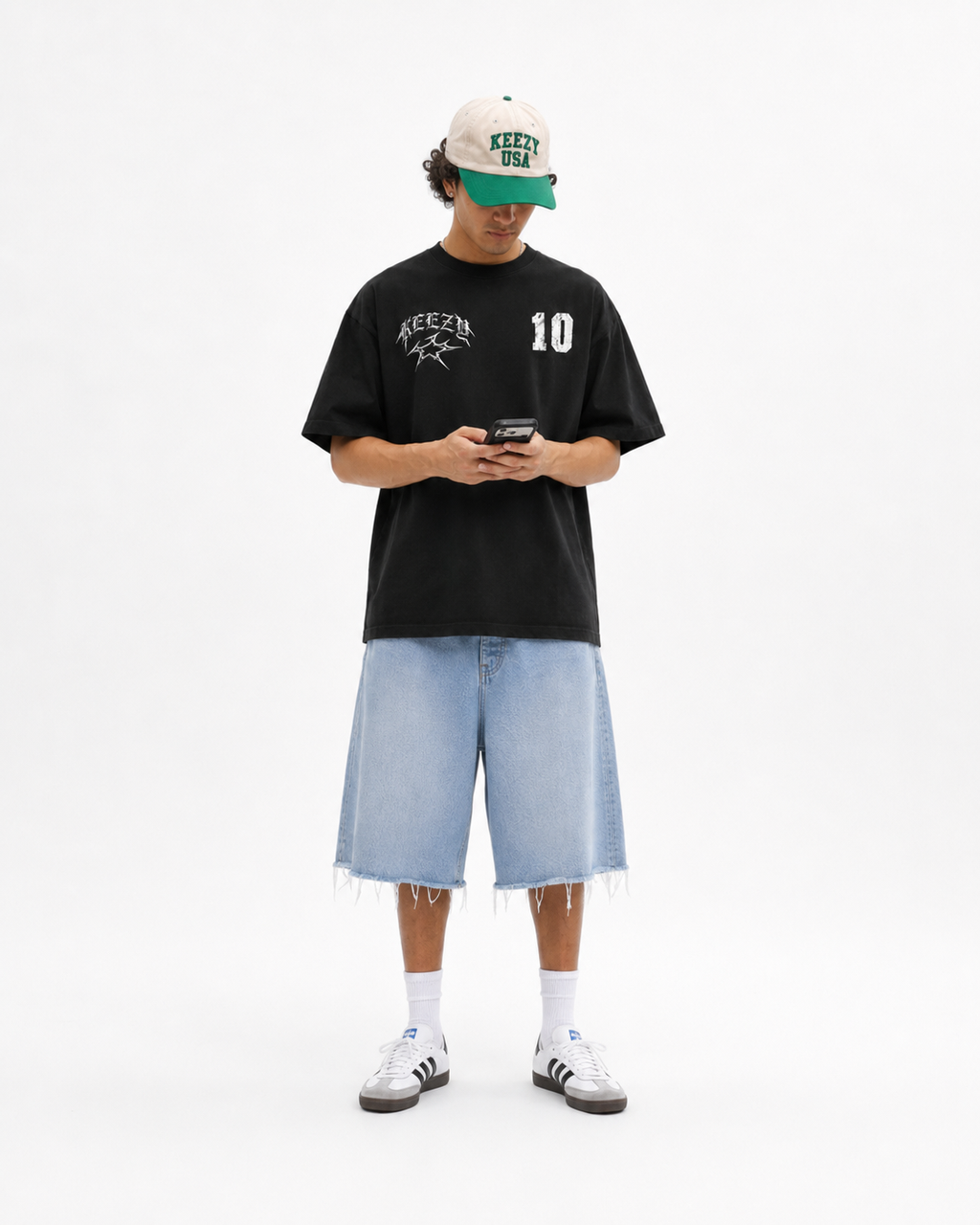 A model stands against a white studio background wearing a black graphic t-shirt with 'KEEZY' and the number '10' on the chest, paired with light-wash denim shorts, white socks, and white sneakers. He is also wearing a beige and green baseball cap and is holding a phone.