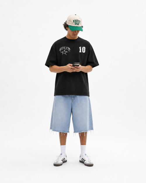 A model stands against a white studio background wearing a black graphic t-shirt with 'KEEZY' and the number '10' on the chest, paired with light-wash denim shorts, white socks, and white sneakers. He is also wearing a beige and green baseball cap and is holding a phone.