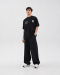 A man modeling a black crewneck t-shirt with white graphic prints on the chest, paired with matching black sweatpants and white sneakers, against a plain light gray studio background.
