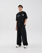 A man modeling a black crewneck t-shirt with white graphic prints on the chest, paired with matching black sweatpants and white sneakers, against a plain light gray studio background.