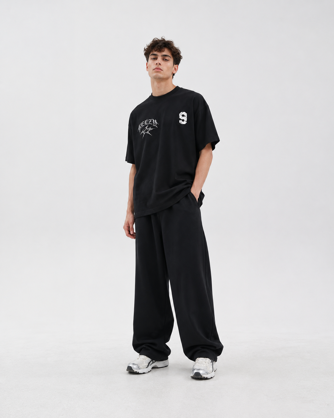 A man modeling a black crewneck t-shirt with white graphic prints on the chest, paired with matching black sweatpants and white sneakers, against a plain light gray studio background.