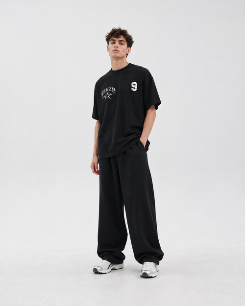 A man modeling a black crewneck t-shirt with white graphic prints on the chest, paired with matching black sweatpants and white sneakers, against a plain light gray studio background.