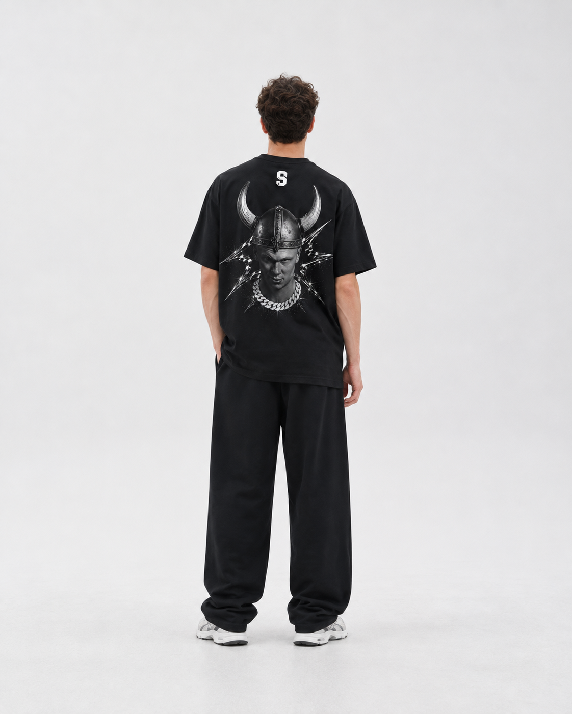 A model viewed from the back wearing a black t-shirt with a large white graphic of a horned HAALAND on the back, paired with black sweatpants and white sneakers, against a light gray studio background. FOOTBALL
