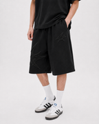 A person wearing black shorts with tonal embroidered graphic details, styled with white crew socks and white sneakers with black stripes, against a light gray studio background.