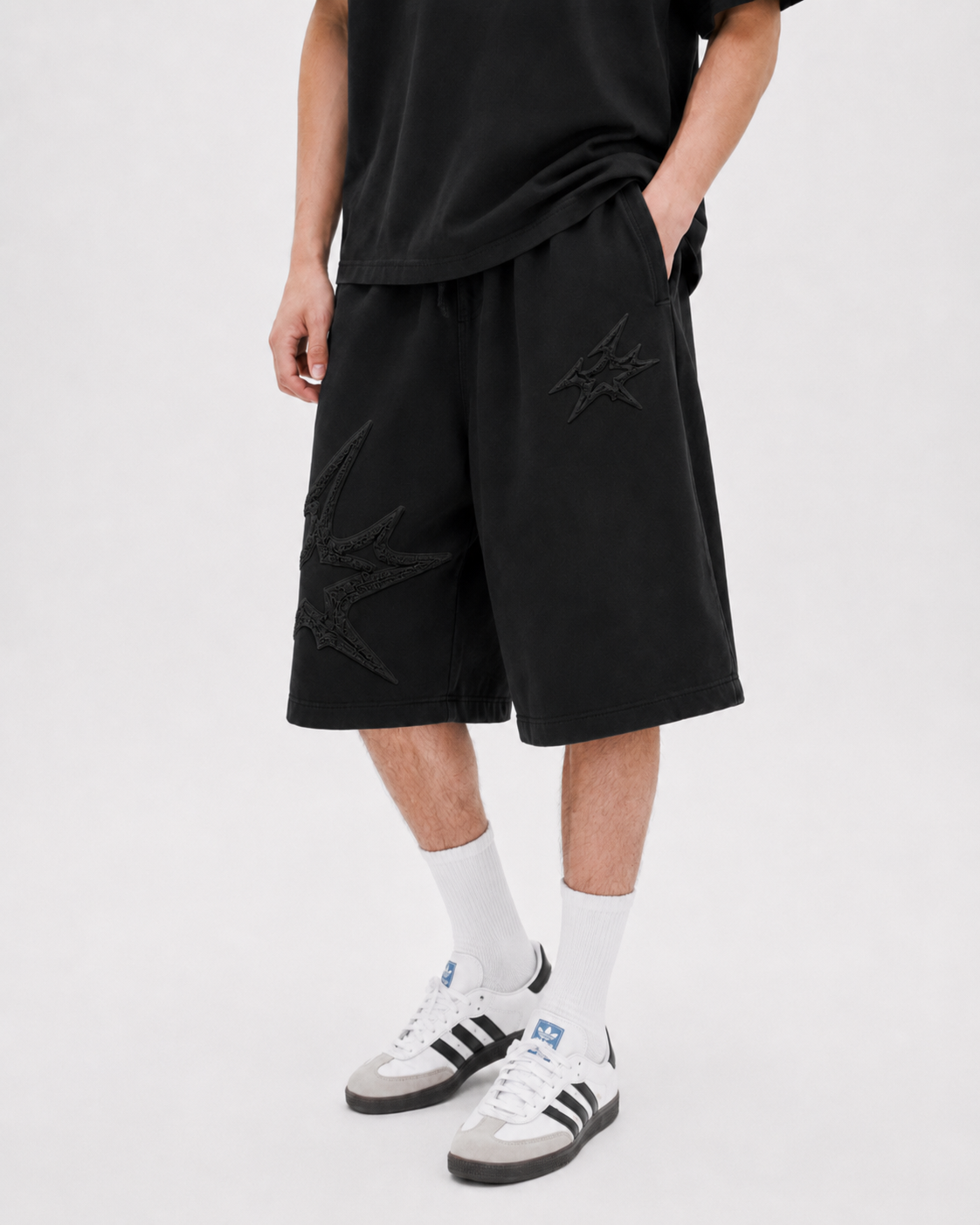 A person wearing black shorts with tonal embroidered graphic details, styled with white crew socks and white sneakers with black stripes, against a light gray studio background.