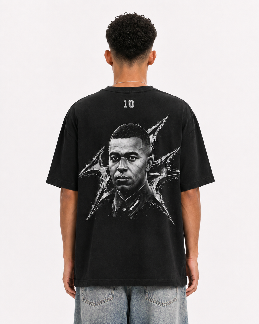 A man viewed from behind wearing a black graphic t-shirt with a large black and white portrait print on the back, paired with light blue jeans, against a plain white background. mbappe