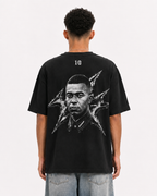 A man viewed from behind wearing a black graphic t-shirt with a large black and white portrait print on the back, paired with light blue jeans, against a plain white background. mbappe