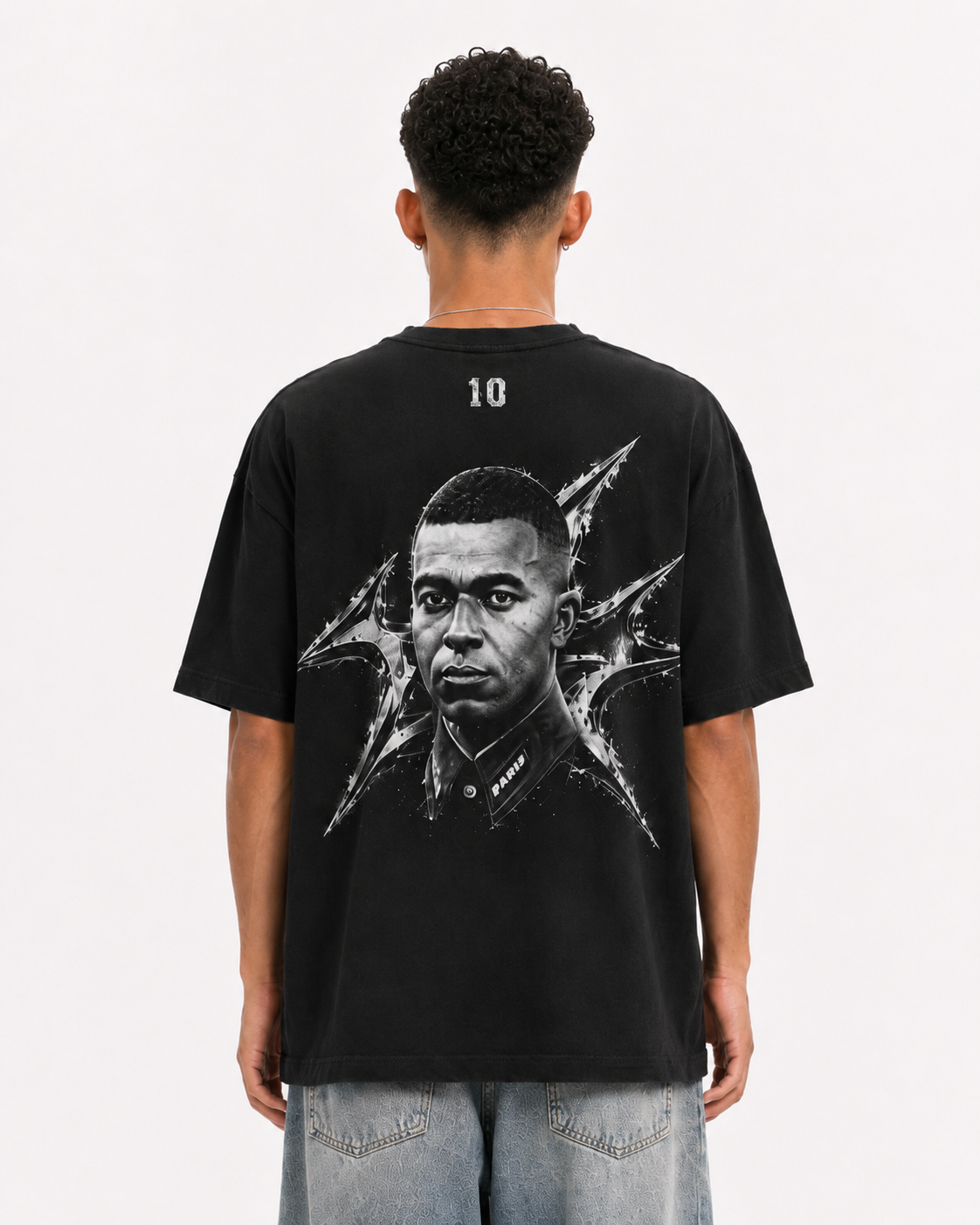 A man viewed from behind wearing a black graphic t-shirt with a large black and white portrait print on the back, paired with light blue jeans, against a plain white background. mbappe