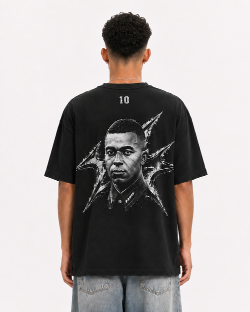 A man viewed from behind wearing a black graphic t-shirt with a large black and white portrait print on the back, paired with light blue jeans, against a plain white background. mbappe