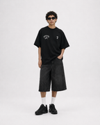 A model stands against a light gray studio background wearing an oversized black graphic T-shirt with white text and a number 7, dark washed denim shorts, white socks, and black sneakers. keezy tshirt 
