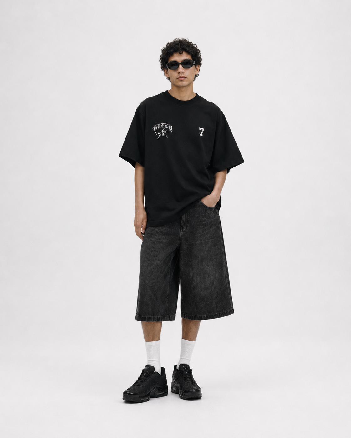 A model stands against a light gray studio background wearing an oversized black graphic T-shirt with white text and a number 7, dark washed denim shorts, white socks, and black sneakers. keezy tshirt 