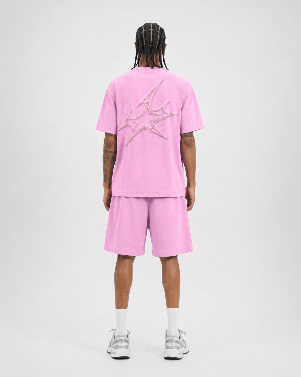 A model shown from the back wearing a pink short-sleeve t-shirt and matching pink shorts, featuring a large metallic silver graphic on the back of the shirt, paired with white socks and silver sneakers against a light gray studio background.