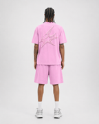 A model shown from the back wearing a pink short-sleeve t-shirt and matching pink shorts, featuring a large metallic silver graphic on the back of the shirt, paired with white socks and silver sneakers against a light gray studio background.