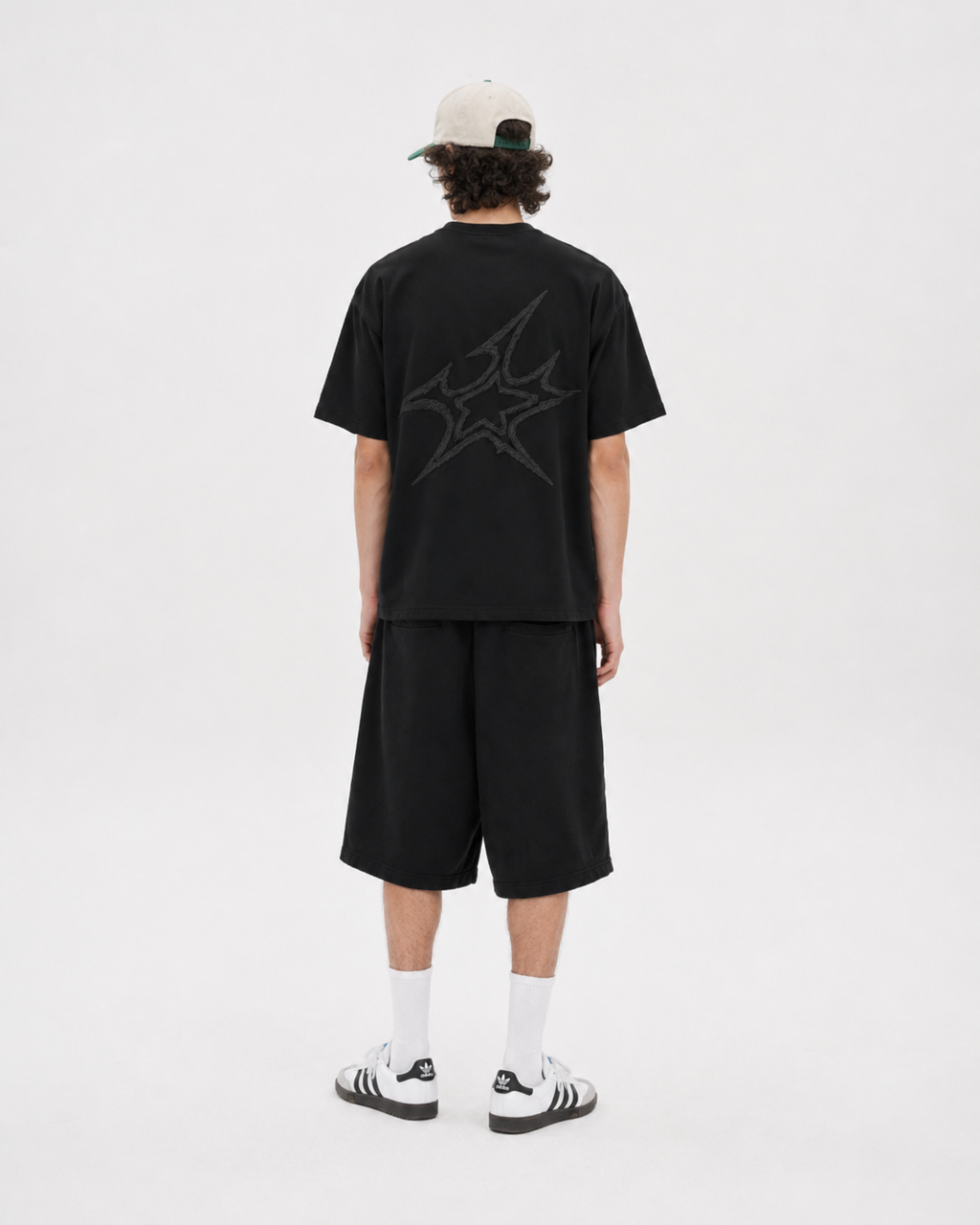 A model viewed from the back wearing a black oversized t-shirt with a dark gray graphic on the back, black shorts, white socks, black and white sneakers, and a beige baseball cap.