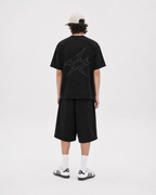 A model viewed from the back wearing a black oversized t-shirt with a dark gray graphic on the back, black shorts, white socks, black and white sneakers, and a beige baseball cap.