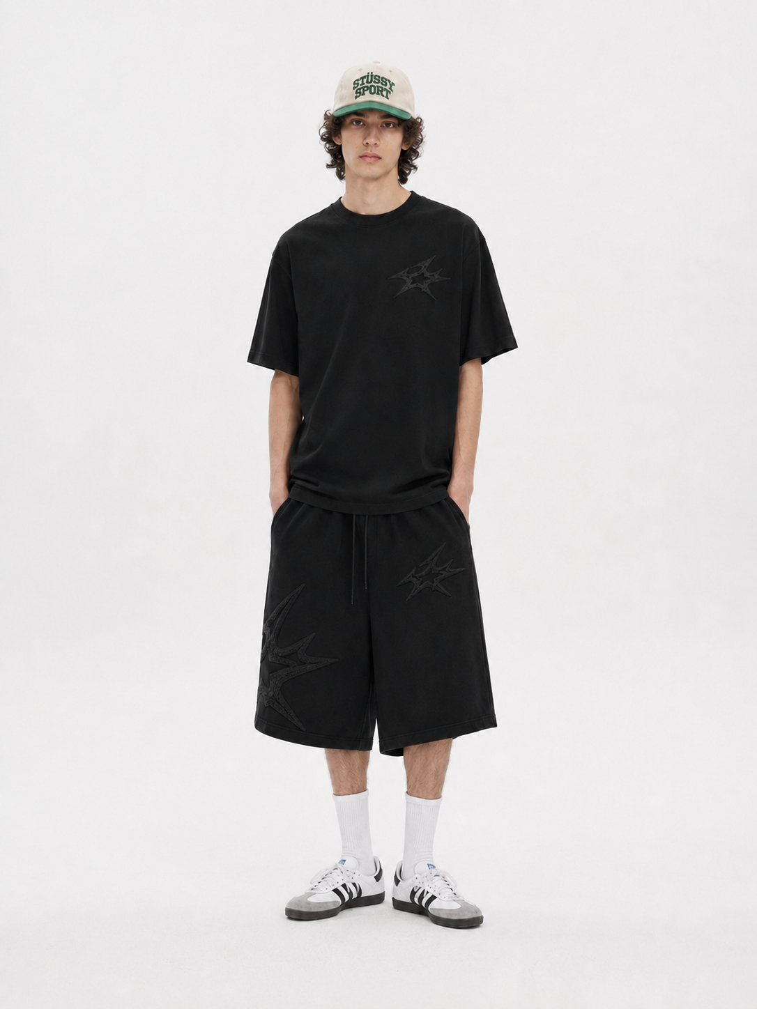 A model stands against a light gray studio background wearing a black short-sleeve t-shirt with a tonal star logo on the chest, matching black shorts, white crew socks, and white sneakers. He is also wearing a beige baseball cap with green lettering.